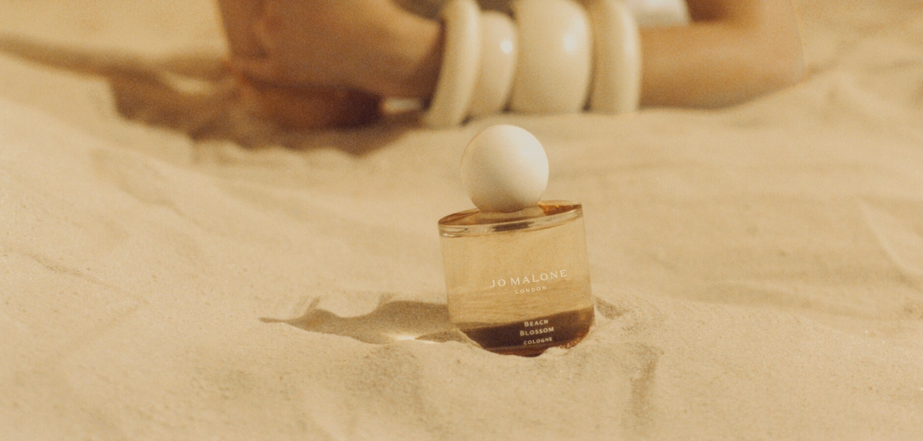 A Jo Malone London cologne sparkles on a beach, with people in beachwear, and holding a cream box
