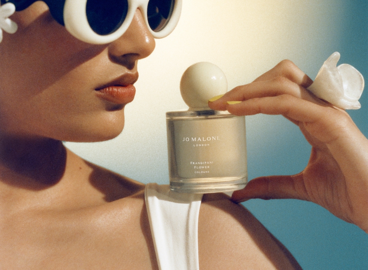 A person wearing white swimwear and sunglasses, holds a Jo Malone London cologne on their shoulder against a blue background. 