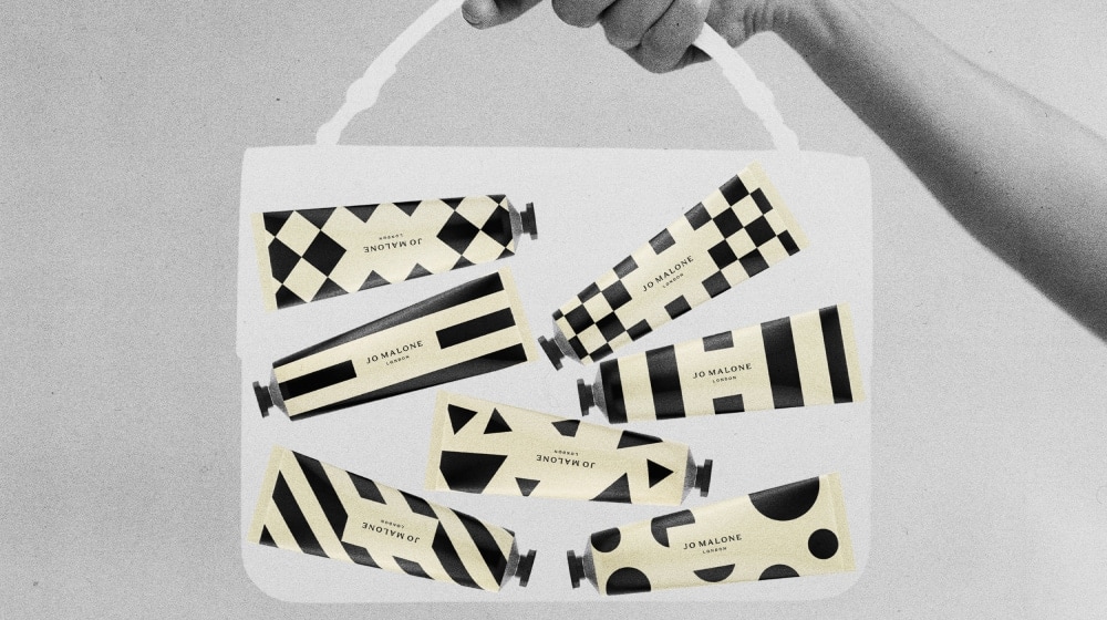 Black, white & cream image of a transparent, medium bag held by a hand, containing seven patterned hand creams in tubes.