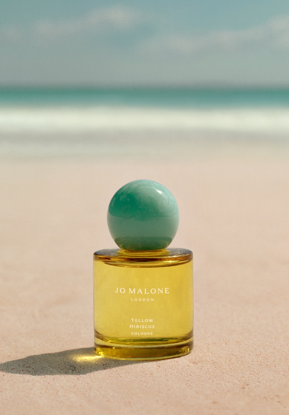 Jo Malone London Yellow Hibiscus cologne sat on a tropical beach. A clear yellow bottle with turquoise cap.