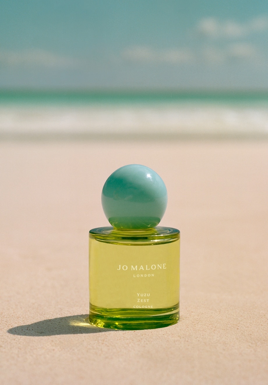 Jo Malone London Yuzu Zest cologne sat on a tropical beach. A clear yellow bottle with turquoise cap.