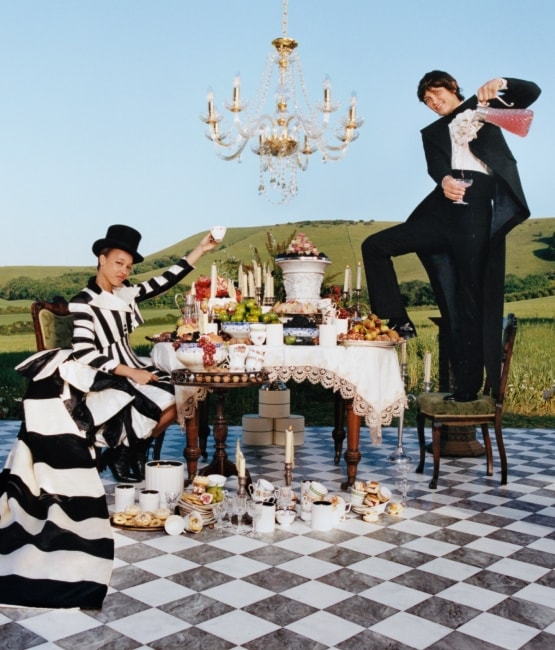 A grand outdoor dining scene with chandeliers, a checkered floor, and an elegantly dressed duo celebrating against rolling hills