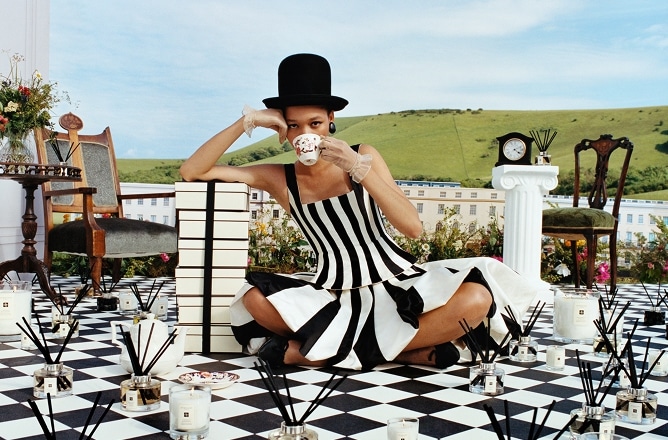 A woman in a striped dress and top hat sips tea on a checkered floor, surrounded by candles, against a countryside backdrop.