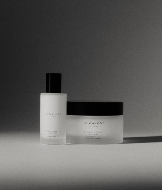 A black and white image of two different Jo Malone London products, one being taller, both with dark lids sit atop a table.