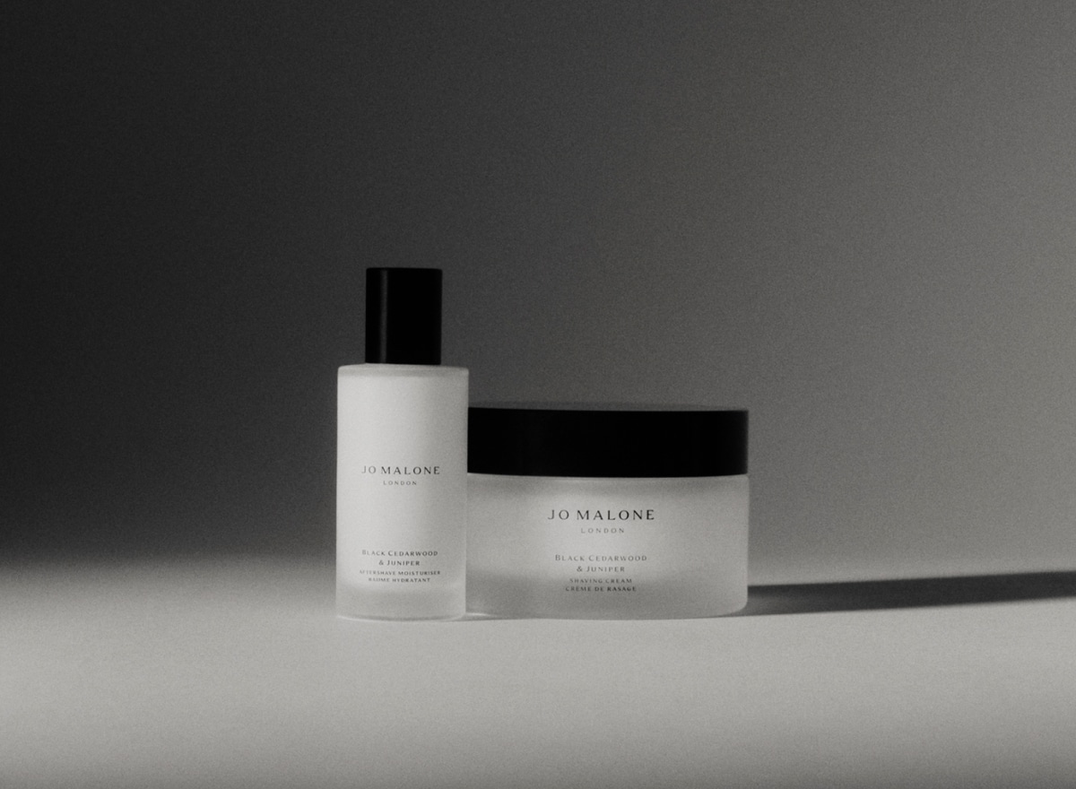 A black and white image of two different Jo Malone London products, one being taller, both with dark lids sit atop a table.