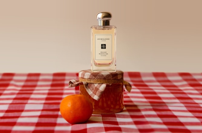 Jo Malone London cologne on a marmalade jar with an orange, styled on a red gingham tablecloth in a playful setup.