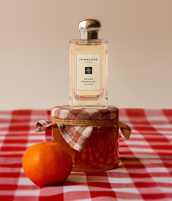 Jo Malone London cologne on a marmalade jar with an orange, styled on a red gingham tablecloth in a playful setup.