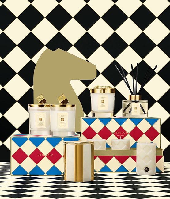 Jo Malone London festive home scents with candles and diffusers in gold-trimmed jars, set on colorful boxes and checkered floor.