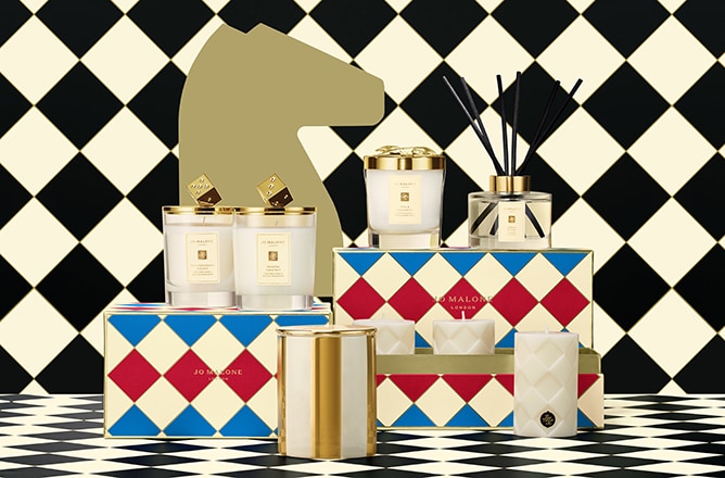 Jo Malone London festive home scents with candles and diffusers in gold-trimmed jars, set on colorful boxes and checkered floor.