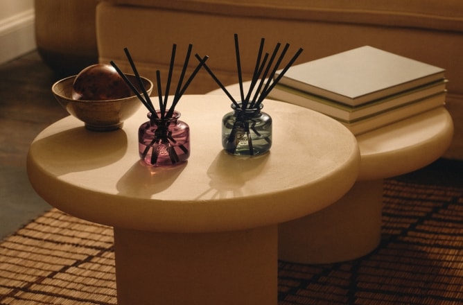 A pair of colourful refillable diffusers on a round table, styled with books and décor in a warm, neutral living room setting.