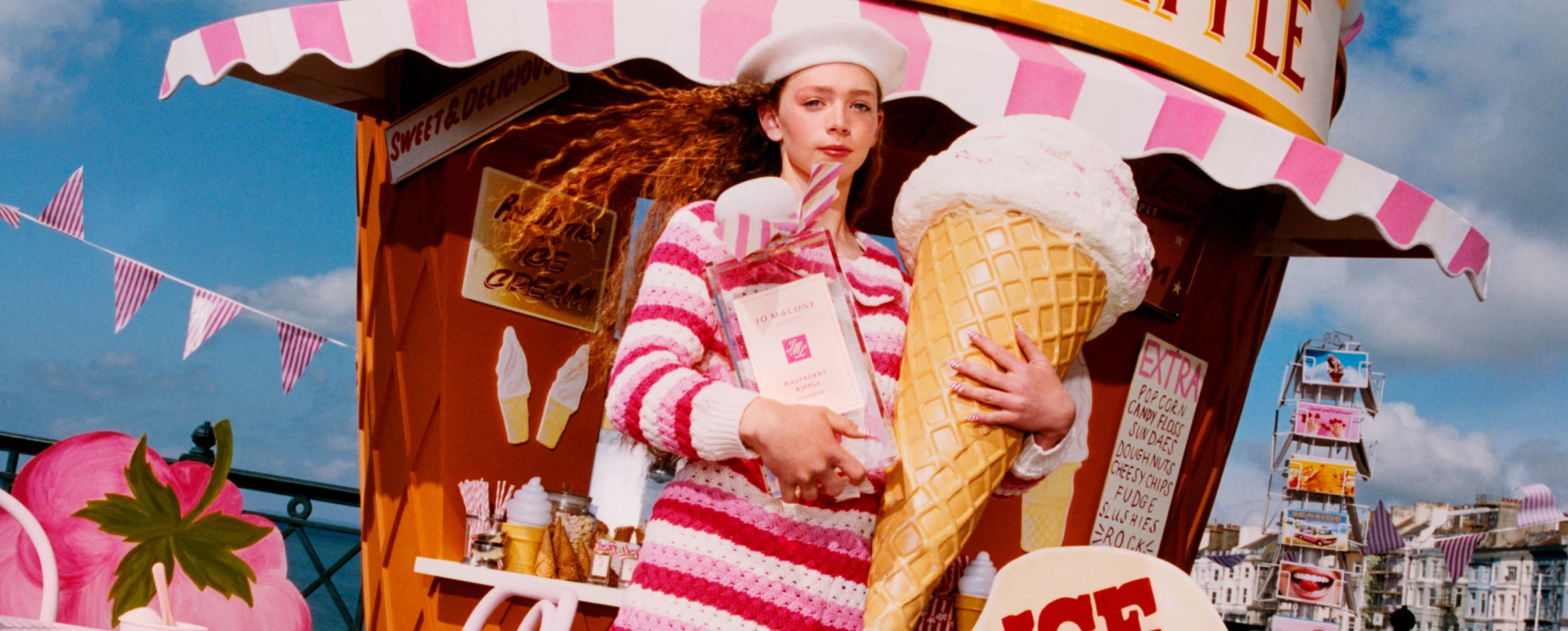 A person in pink takes an icecream then walks along a pier containing vibrant beach props before dropping the icecream. 