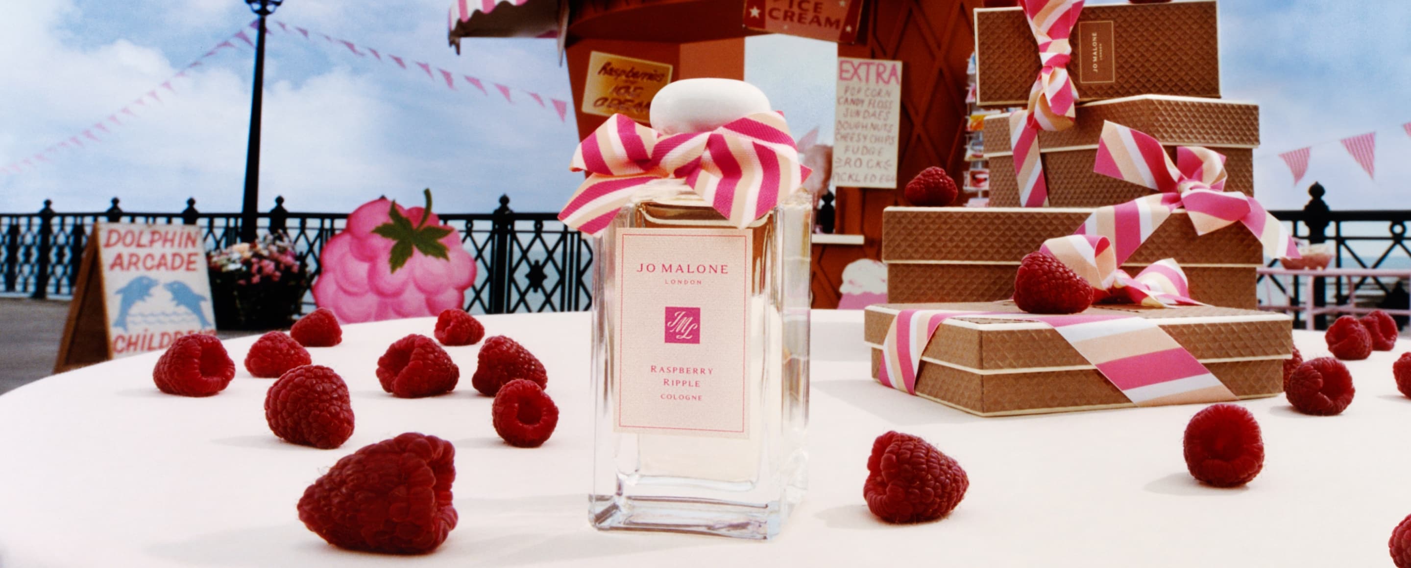 A Jo Malone London cologne with a pink bow surrounded by raspberries and a giant cone hut next to stacked waffle gift boxes.