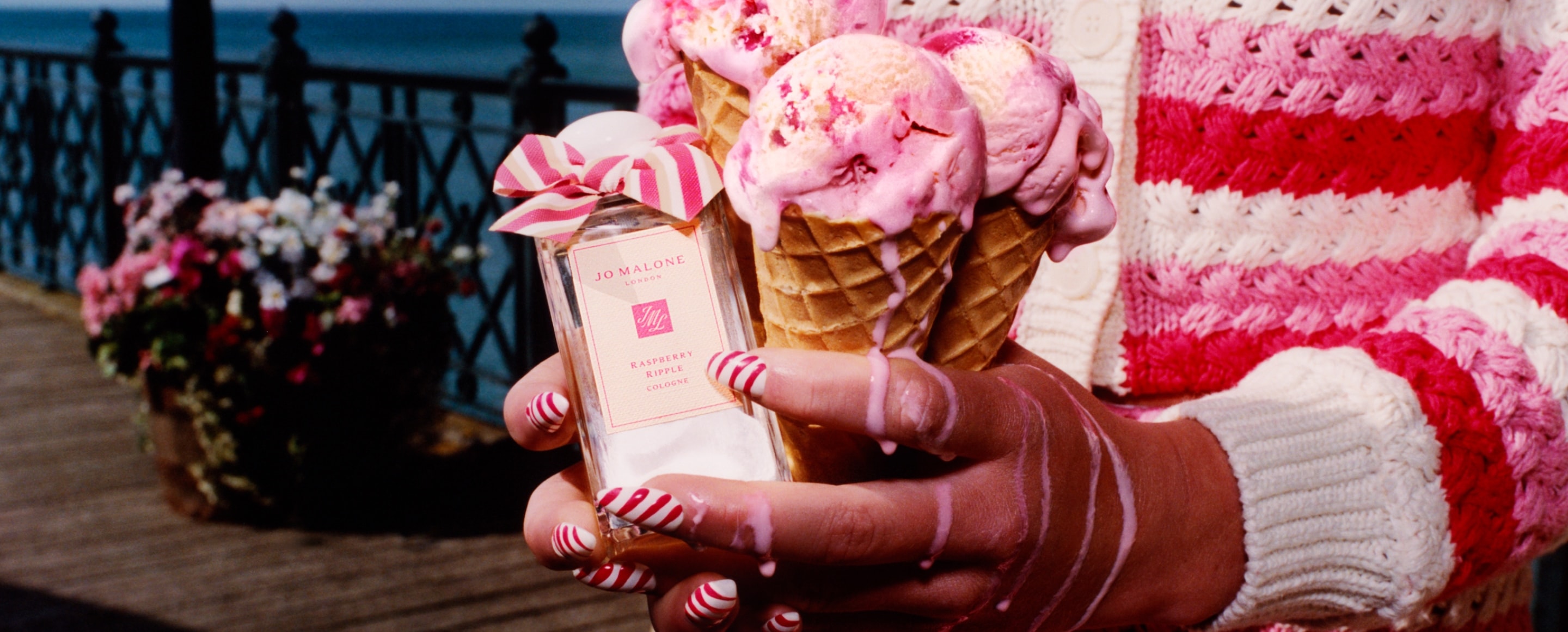 Three ice cream cones held next to a Jo Malone London cologne adorned with a pink bow, set against a scenic waterfront pier.