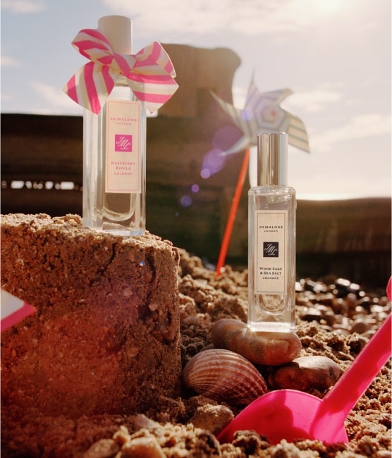Various Jo Malone London products sit atop a stoney beach with a towel, pink bucket, vanity case and shells. 