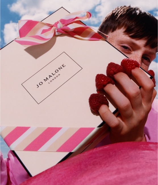 A Jo Malone London box with a pink striped bow is held by a person who has raspberries on the tips of their fingers