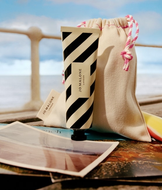 A stripey hand cream tube sits atop a wooden bench on a pierside with a coloured edible rock, vanity pouch and postcard. 