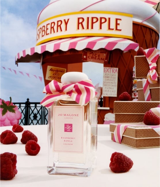 A Jo Malone London cologne with a pink bow surrounded by raspberries and a giant cone hut next to stacked waffle gift boxes.