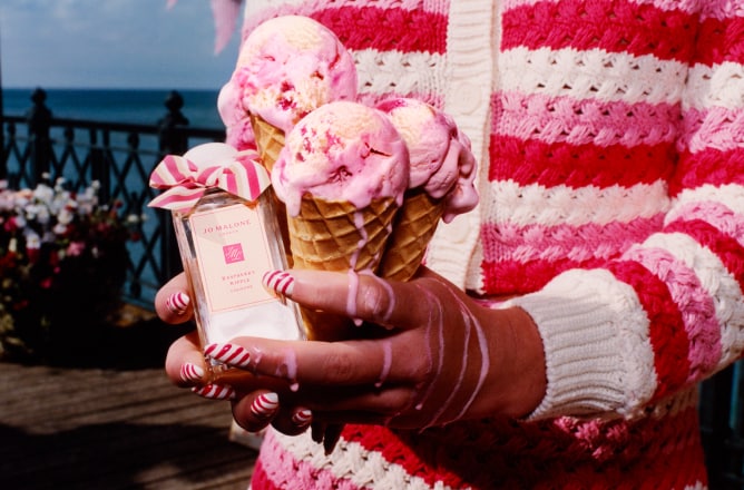 Three ice cream cones held next to a Jo Malone London cologne adorned with a pink bow, set against a scenic waterfront pier.
