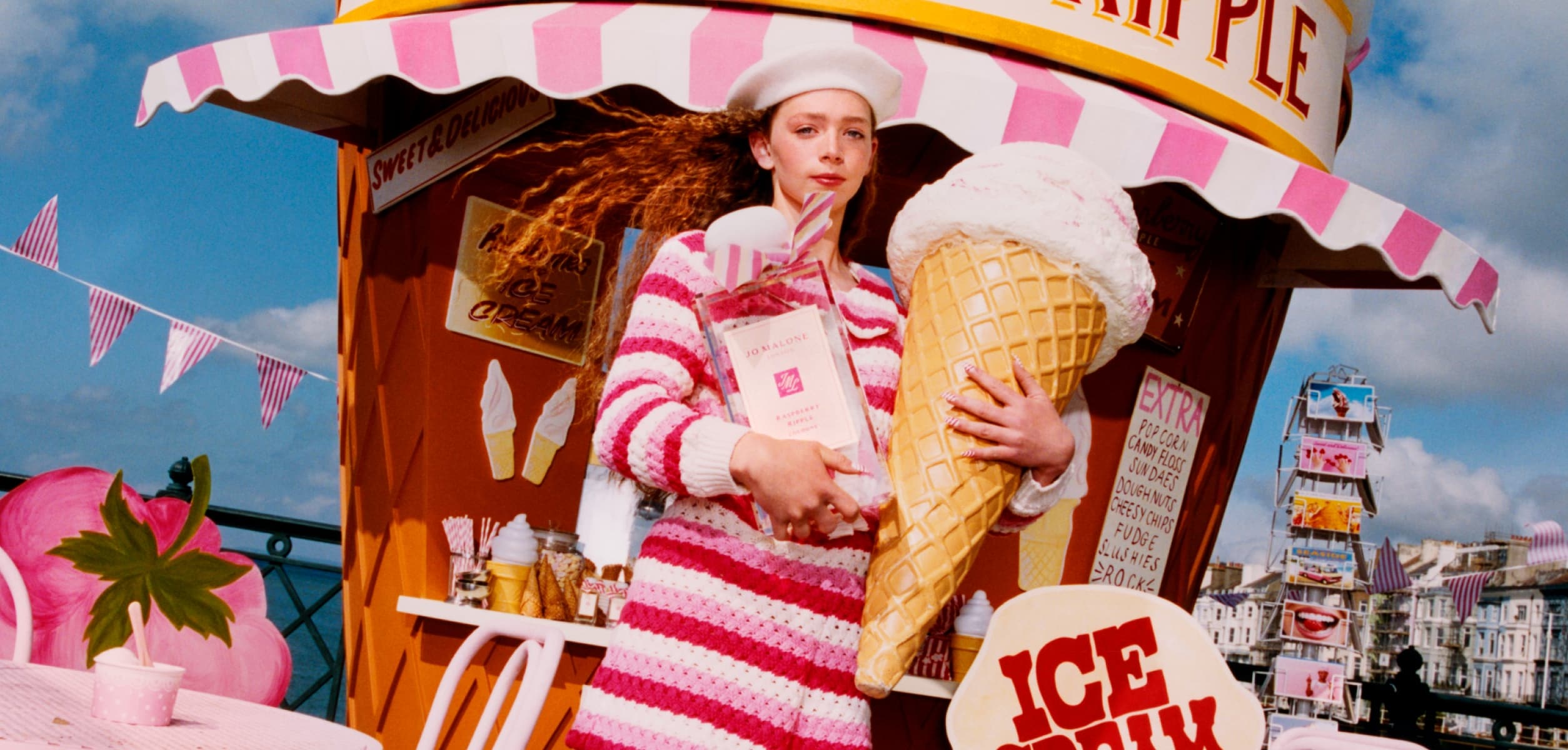 A person in pink holds an oversized ice cream and Jo Malone London cologne on a pier, with pink seating, and a giant cone hut.