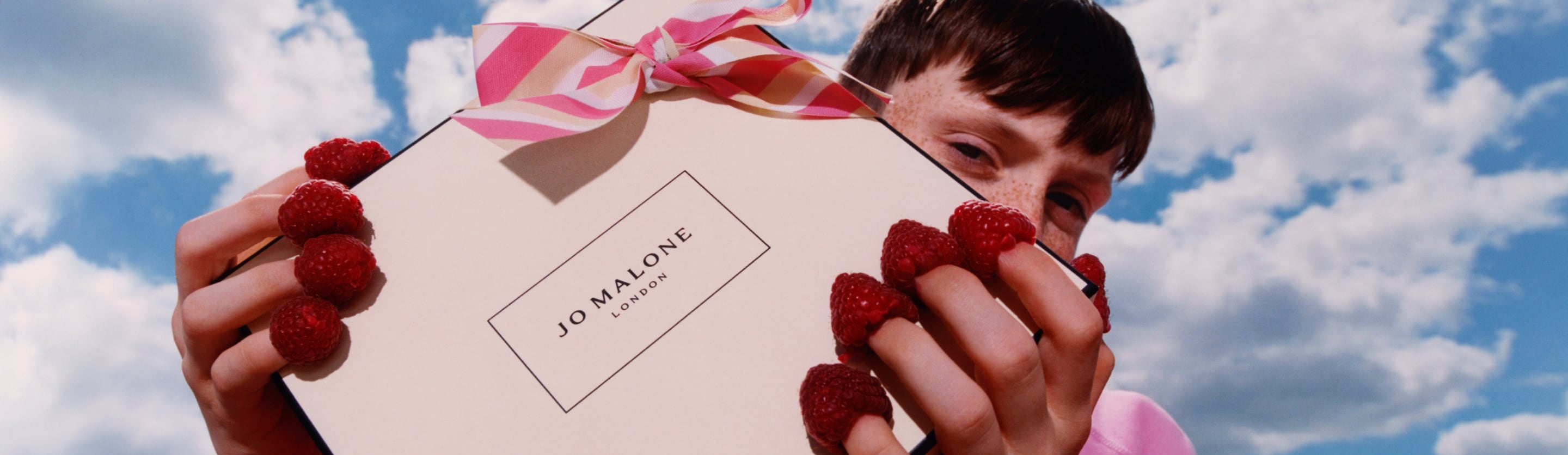 A Jo Malone London box with a pink striped bow is held by a person who has raspberries on the tips of their fingers, against a b