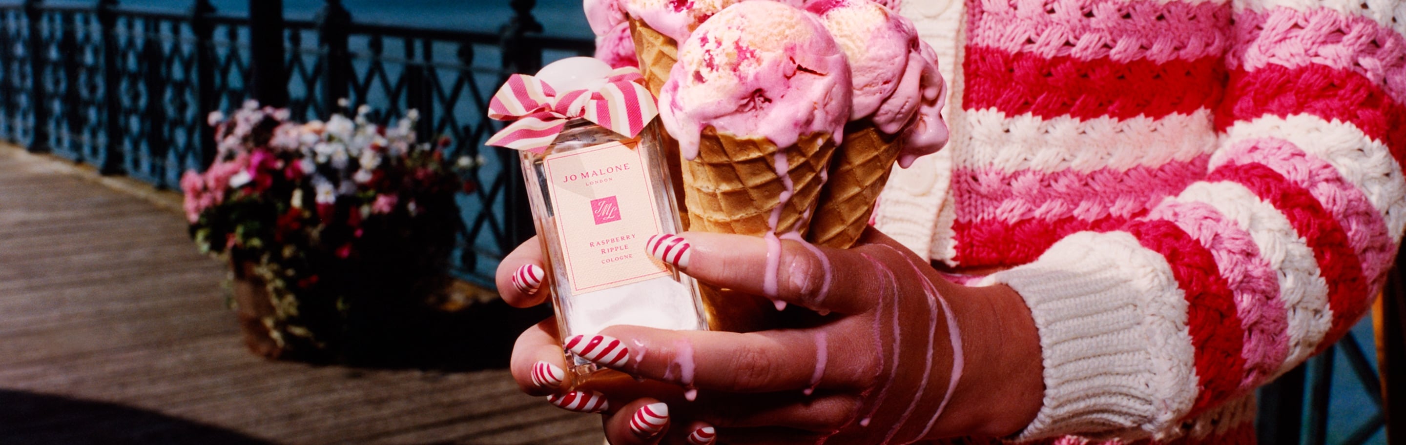 Three ice cream cones held next to a Jo Malone London cologne adorned with a pink bow, set against a scenic waterfront pier.