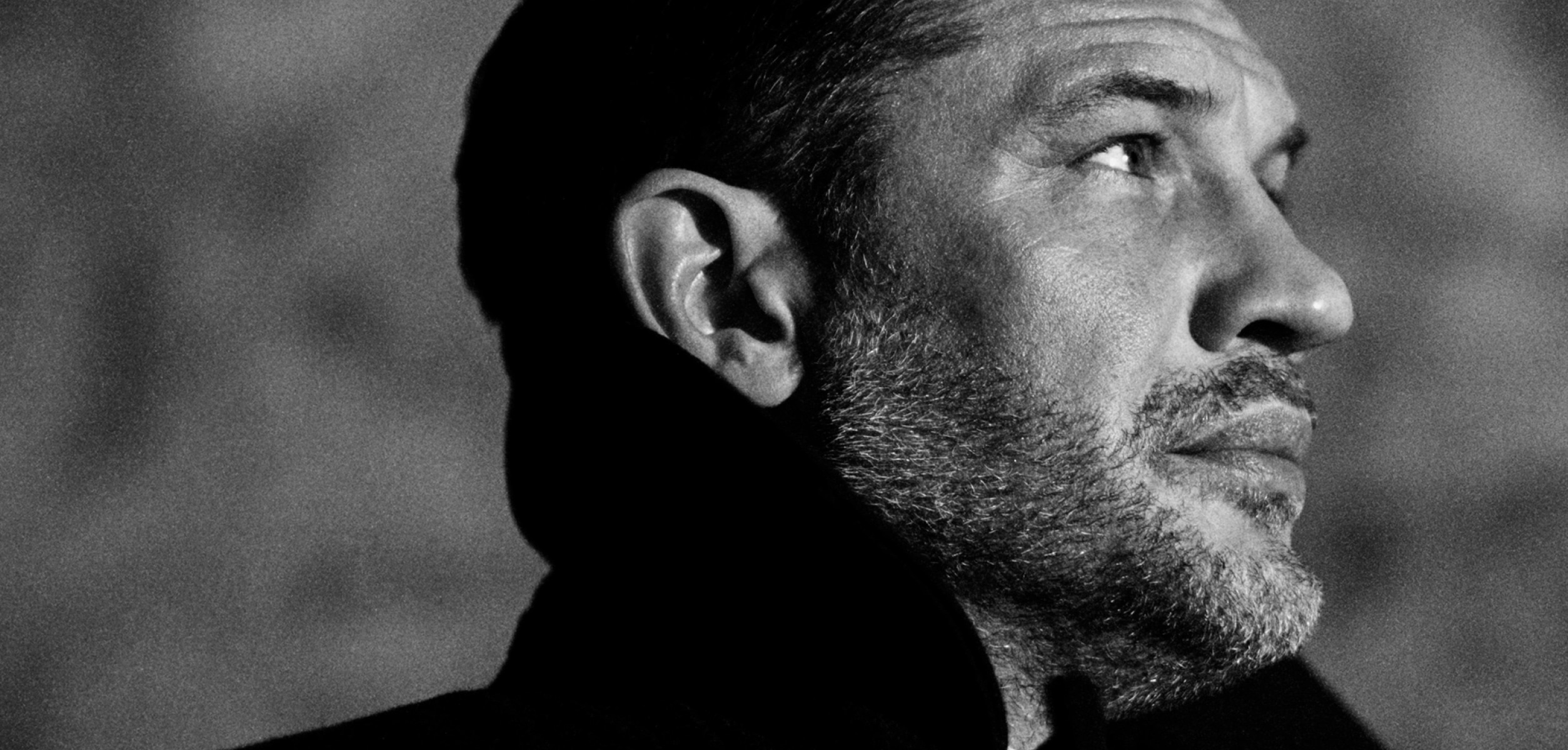 'A side portrait of Tom Hardy in black and white, gazing into the distance. He wears a formal coat, with an upturned collar.