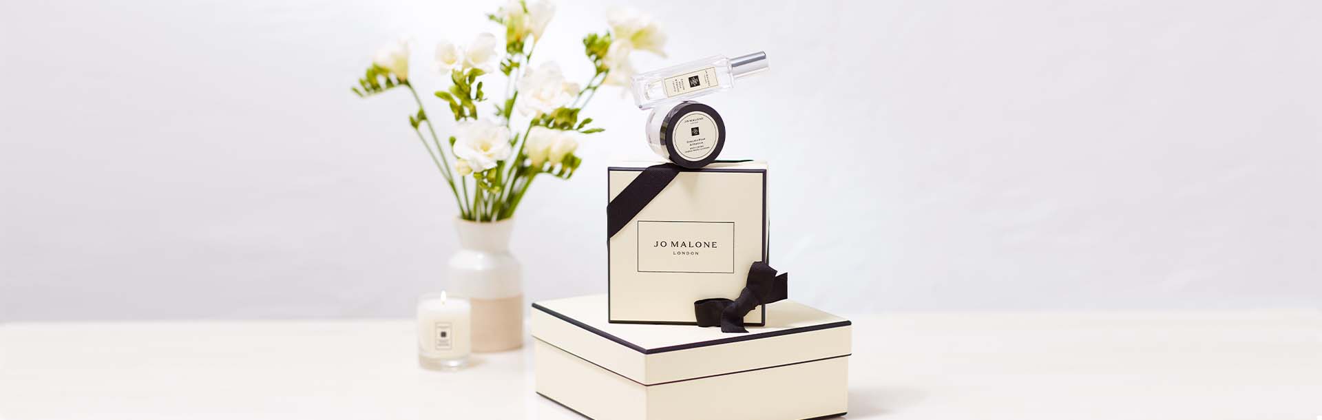image of jo malone cream and black boxes stacked with a body creme balanced on top and flowers in a vase behind