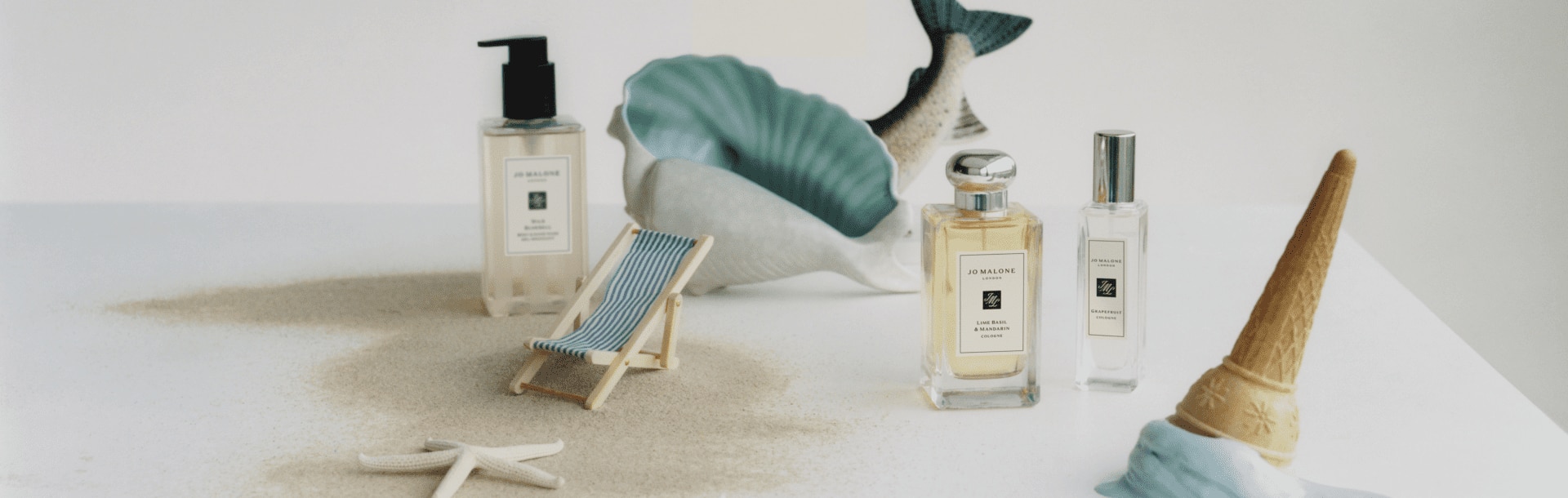 image of jo malone london wild bluebell body & hand wash next to two colognes with a mini deckchair & sandy background