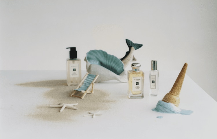 image of jo malone london wild bluebell body & hand wash next to two colognes with a mini deckchair & sandy background